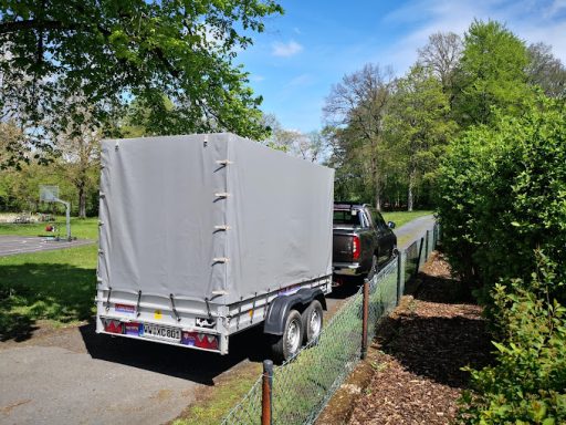 Ein Anhänger mit grauem Planenaufbau parkt an einer Straße, umgeben von Bäumen.