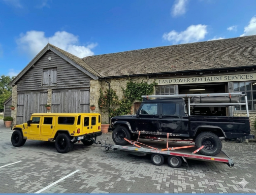 Gelber Hummer neben einem schwarzen Geländewagen auf einem Anhänger vor einer Scheune.